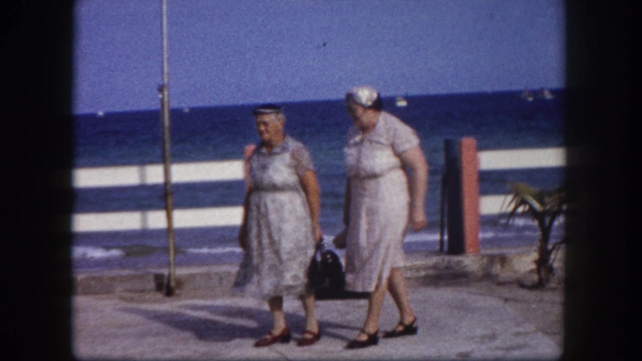 Two women in 1960s summer dresses walk a seafront promenade, ocean horizon and palm tree visible, circa 1960 Florida, filmed on 8mm.