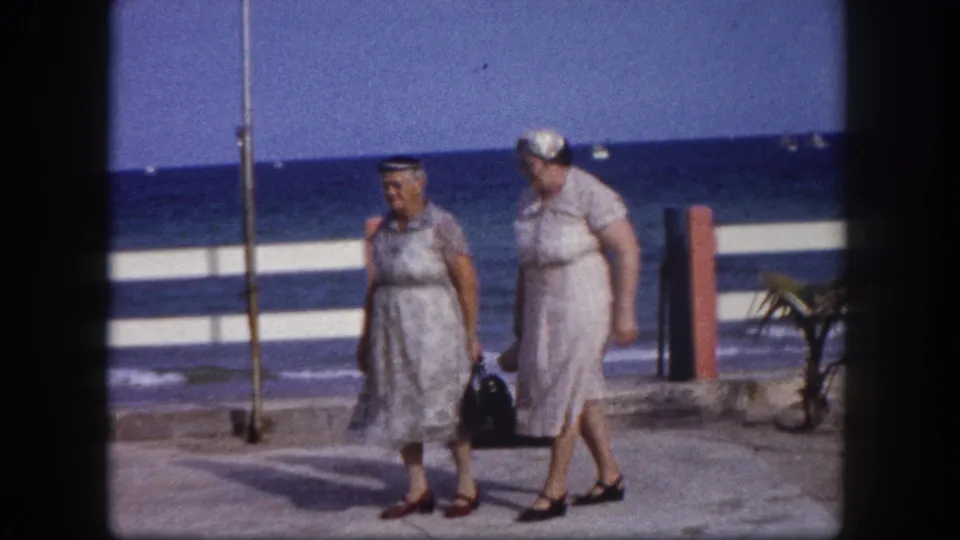 Two women in 1960s summer dresses walk a seafront promenade, ocean horizon and palm tree visible, circa 1960 Florida, filmed on 8mm.