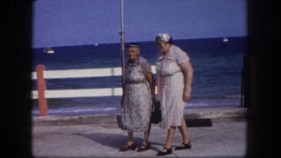 Two women in early-1960s summer attire walk a Florida seafront promenade; striped barrier rail and blue sea horizon visible in background.
