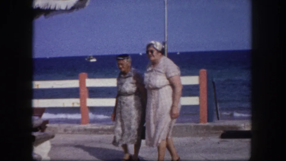 Two women in 1960s summer attire walk past red bollards on a Florida seafront promenade; small boats visible on the ocean horizon.