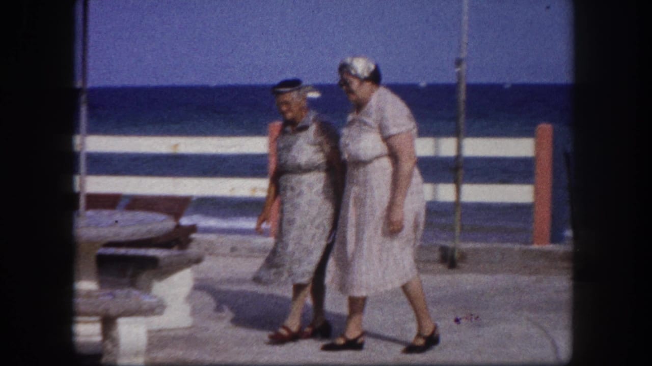 Two older women in early-1960s day dresses and small hats walking along a Florida coastal promenade, stone pedestal picnic tables and blue ocean visible behind a striped barrier, circa 1960.