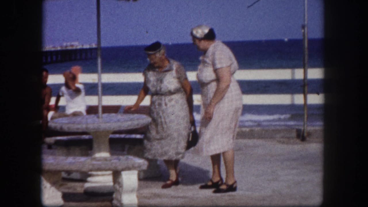 Two older women in matching pale dresses pausing near a stone pedestal table at a Florida coastal pier, seated beachgoers and open ocean visible behind, circa 1960.