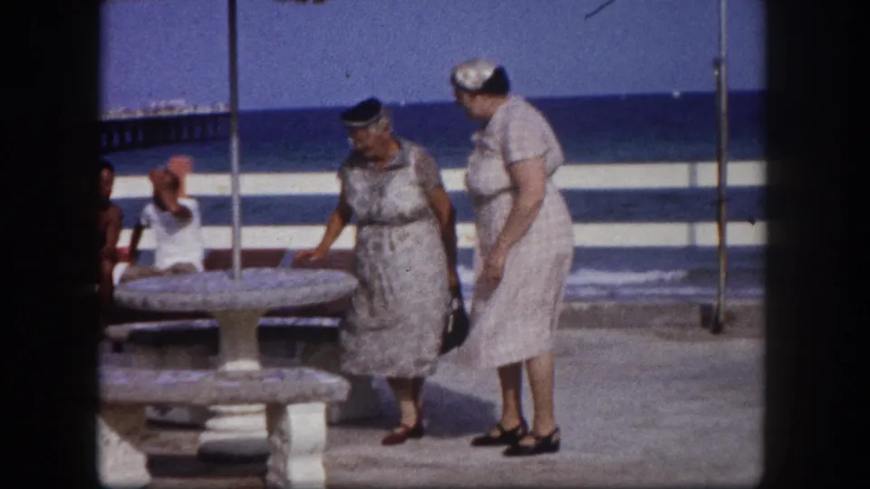 Two older women in matching pale dresses pausing near a stone pedestal table at a Florida coastal pier, seated beachgoers and open ocean visible behind, circa 1960.