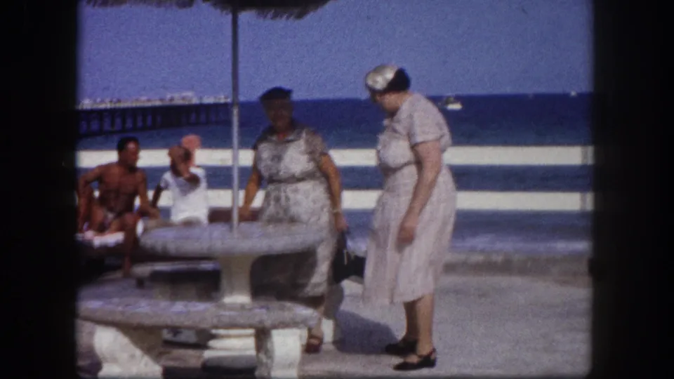 A woman in a pale dress bends toward a stone pedestal table on a Florida coastal promenade; seated background figures relax in the sun nearby, ocean visible behind, circa 1960.