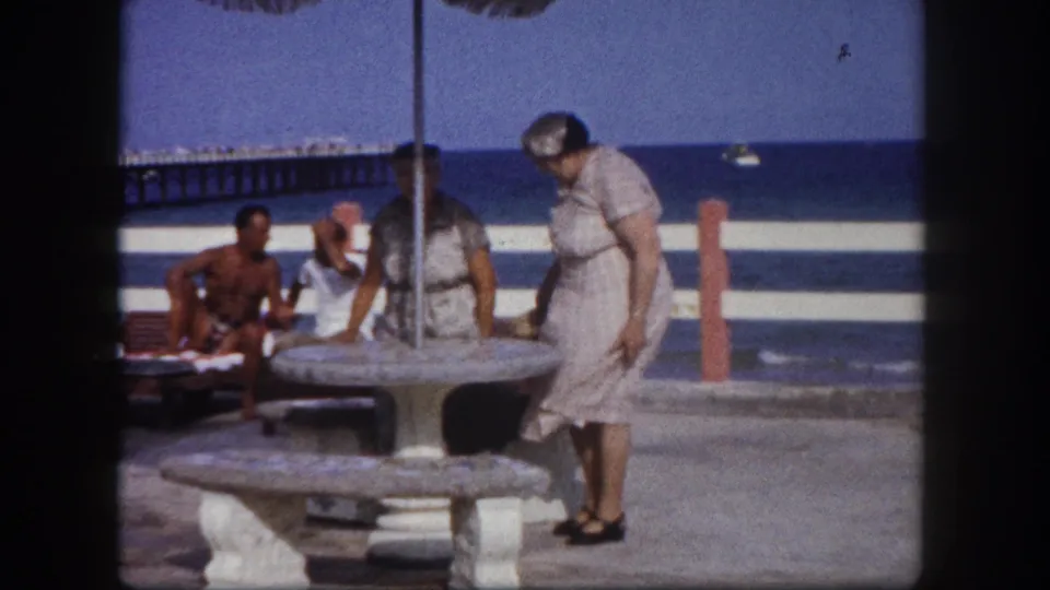 A woman in a pale mid-century dress leans over a stone outdoor table at a Florida coastal pier, seated beachgoers and ocean behind orange-red bollard posts, circa 1960.
