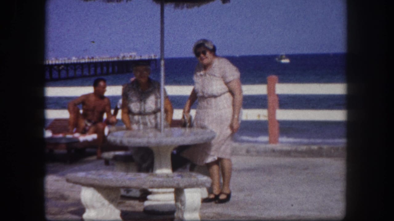 Older woman in a light cotton dress standing at a round concrete pedestal picnic table under a thatched canopy; ocean and wooden pier visible behind, Tampa, Florida, circa 1960.