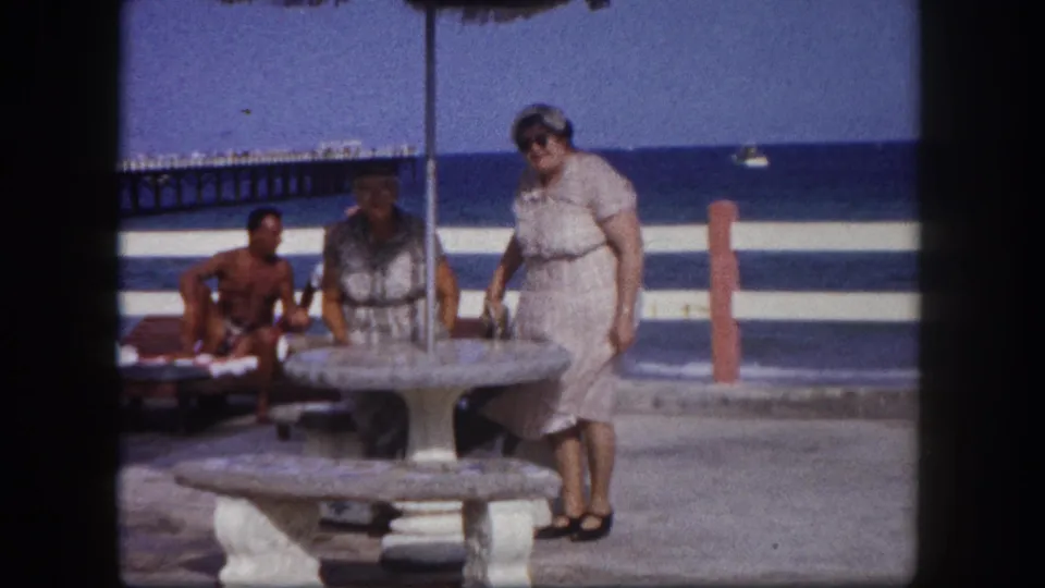 Older woman in a light cotton dress standing at a round concrete pedestal picnic table under a thatched canopy; ocean and wooden pier visible behind, Tampa, Florida, circa 1960.