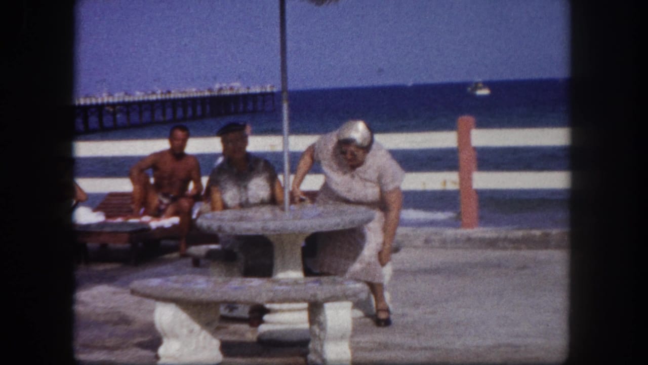 Older woman leaning over a round concrete beach table, second light-haired figure visible at right, shirtless man seated behind with ocean and pier beyond.
