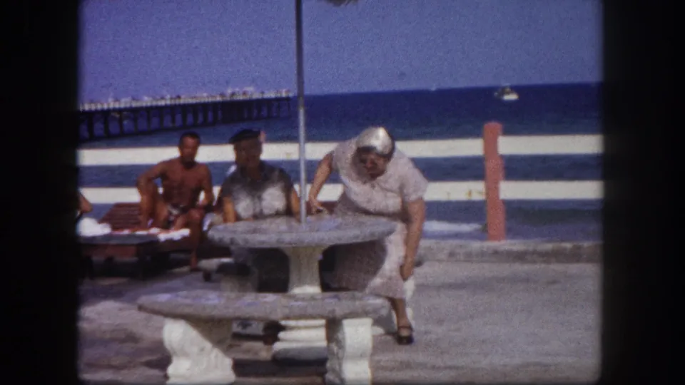 Older woman bending over a round concrete pedestal table, a second light-haired figure at right, shirtless man in background, wooden pier over blue ocean; Tampa, Florida, circa 1960.