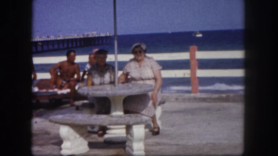Older woman at a round concrete pedestal table under a thatched canopy; two figures in swim trunks seated in background with ocean beyond; Tampa, Florida, circa 1960.