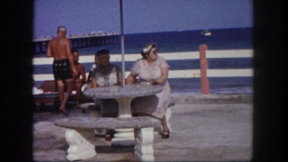 Older woman and companion at a round concrete pedestal beach table, shirtless man in dark swim trunks in middle ground, wooden pier and blue ocean behind; Tampa, Florida, circa 1960.