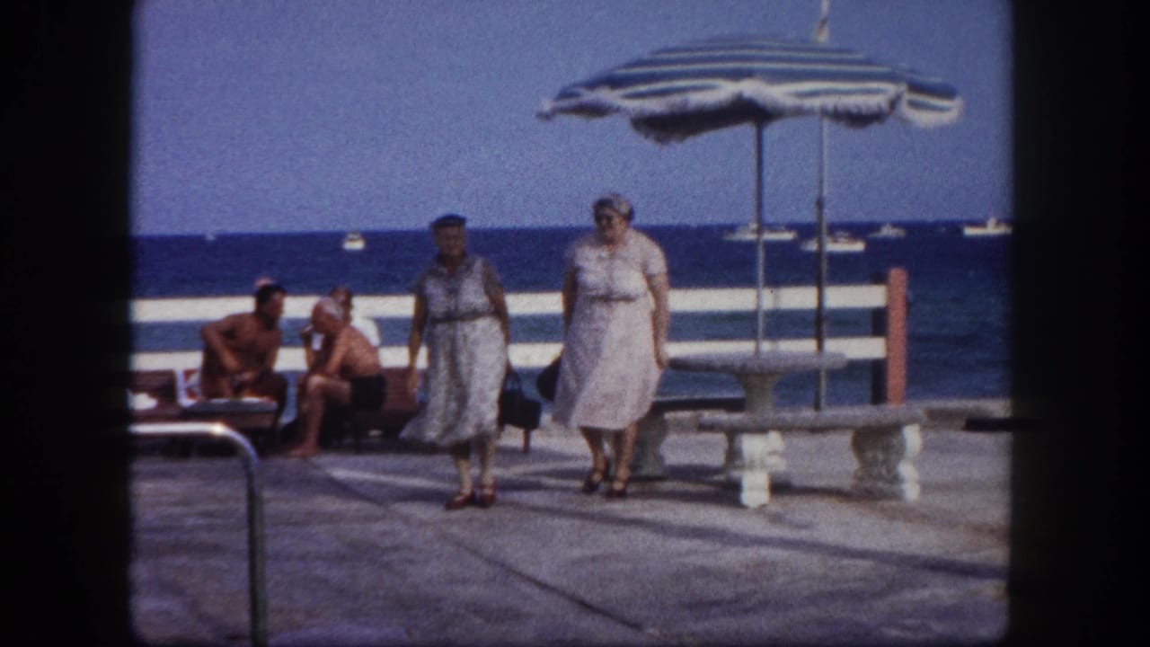 Two elderly women in light belted dresses walk toward camera on a paved oceanfront terrace; striped beach umbrella and men in swimwear visible behind them.