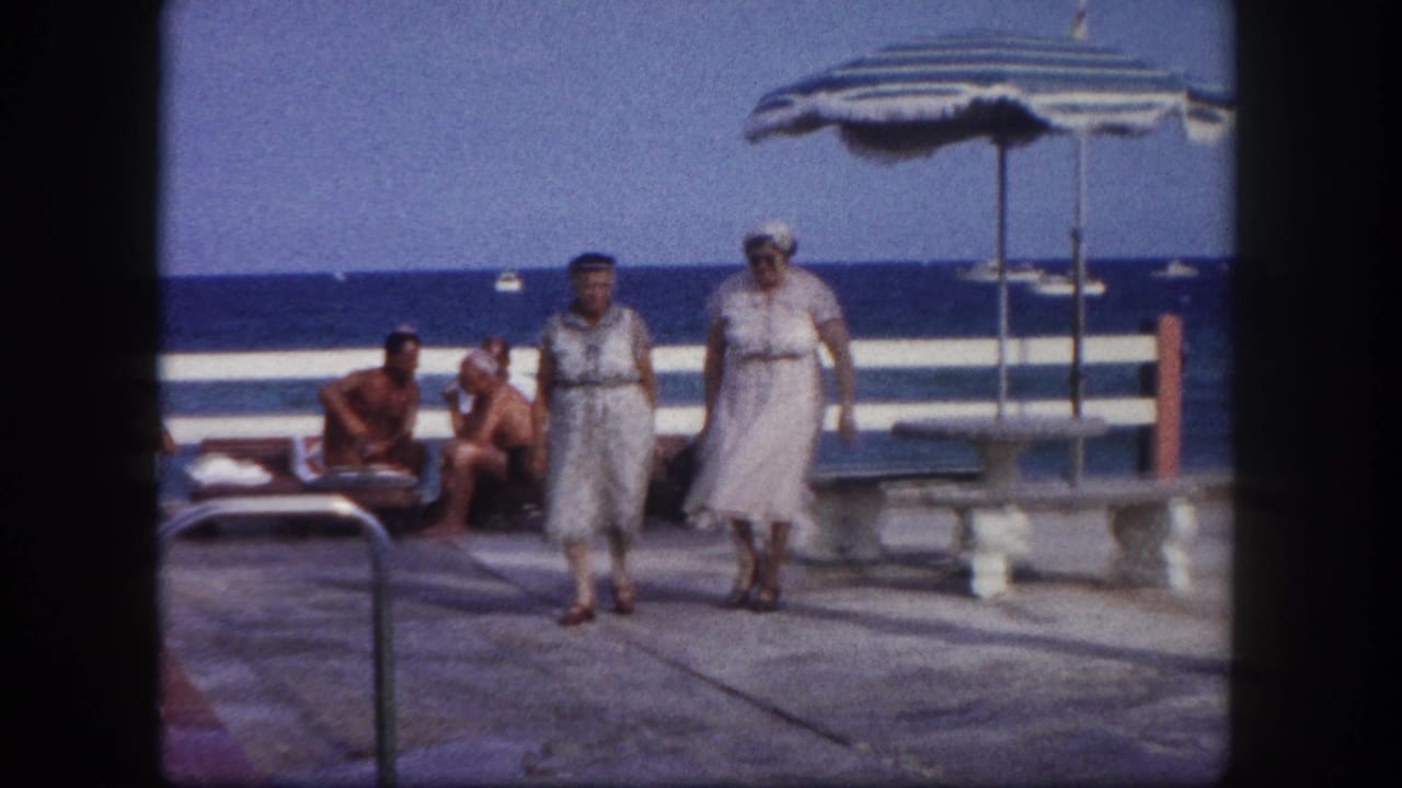 Two elderly women in light dresses walking closer toward camera; striped umbrella at upper right, seated man in swim trunks visible behind them.