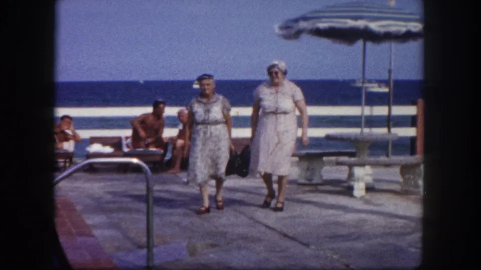 Mid-shot of two elderly women in belted dresses crossing a paved beachside terrace; two striped umbrellas and lounging sunbathers visible, Tampa, Florida area, circa 1960.