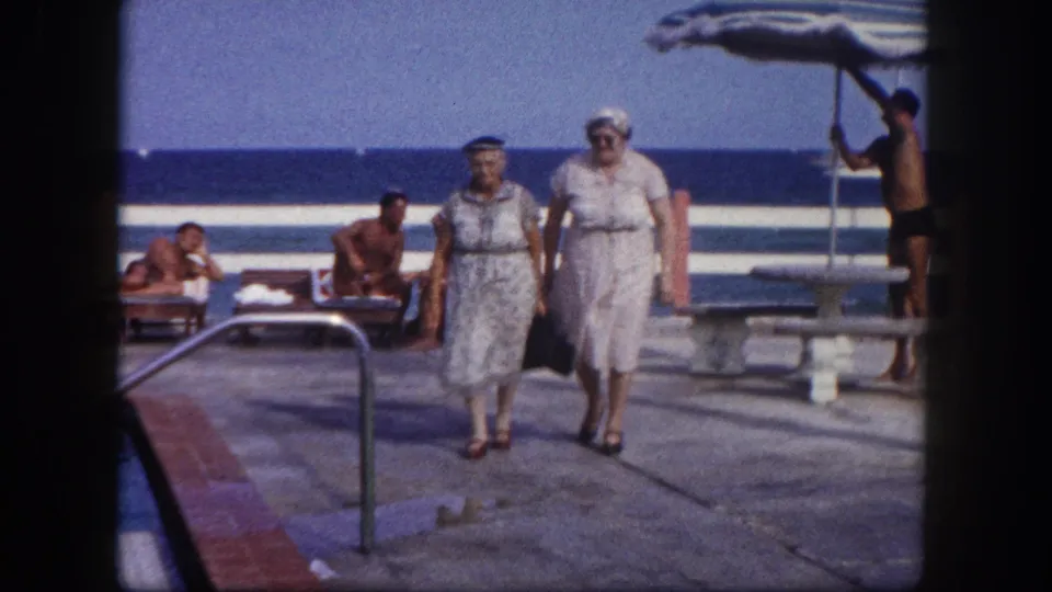 Close-approach shot of two elderly women in belted dresses and hats on a paved coastal terrace; sunbathers and a striped umbrella visible, consistent with a 1960s Florida beach facility.