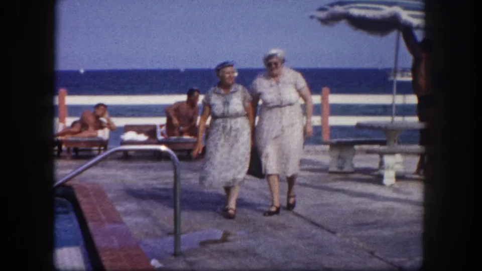 Two older women in early 1960s resort attire walk toward camera on a concrete pool deck at what appears to be a Florida oceanfront resort, circa 1960.