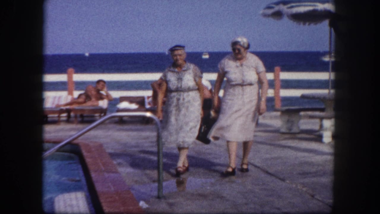 Two older women in pale dresses and hats walking along a resort pool deck, ocean horizon and reclining guests visible behind them.