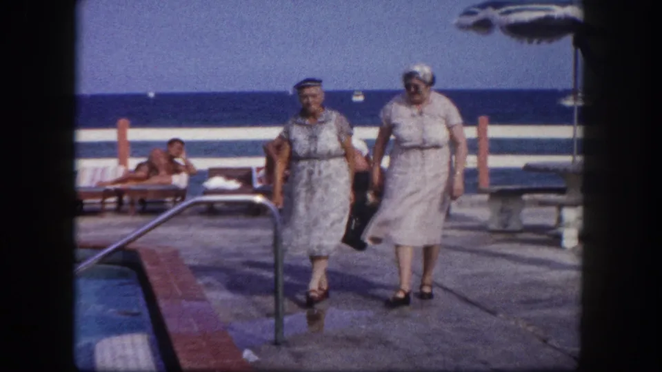 Two older women in mid-century resort wear walk toward camera on a concrete pool deck; ocean and reclining guests on lounge chairs visible in the background, circa 1960.