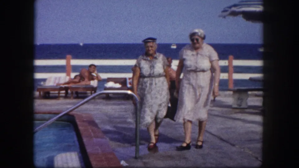 Closer view of two older women in early 1960s resort attire on a pool deck; metal pool handrail visible at right with ocean and guests in the background, circa 1960.