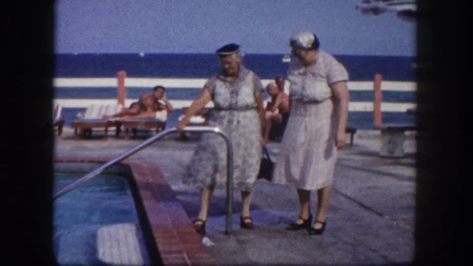 Two older women near the pool handrail on what appears to be a Florida oceanfront resort deck; one reaches toward the rail as reclining guests and blue ocean appear in the background, circa 1960.