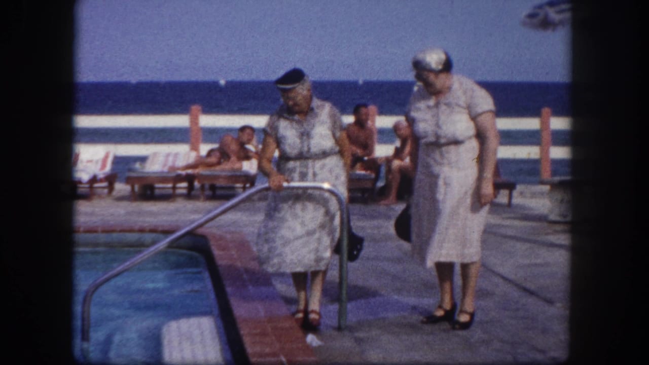Two women in early-1960s summer attire at a Florida beach resort pool; one grips a metal handrail at the pool entry while the other stands nearby; ocean and lounge chairs visible behind a white fence
