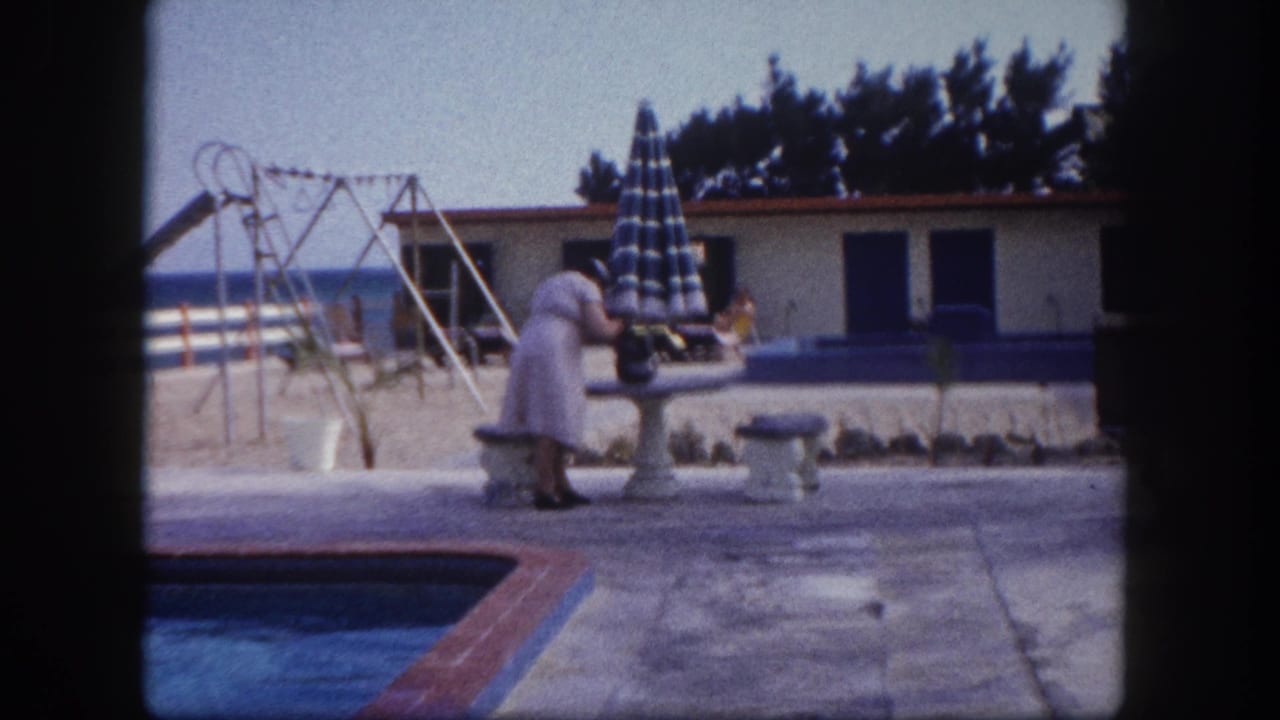 A woman in a light 1960s dress stands near a concrete pedestal beside a Florida beach resort pool; a furled striped umbrella, swing-set frame, low white building with blue shutters, and ocean are visible behind her