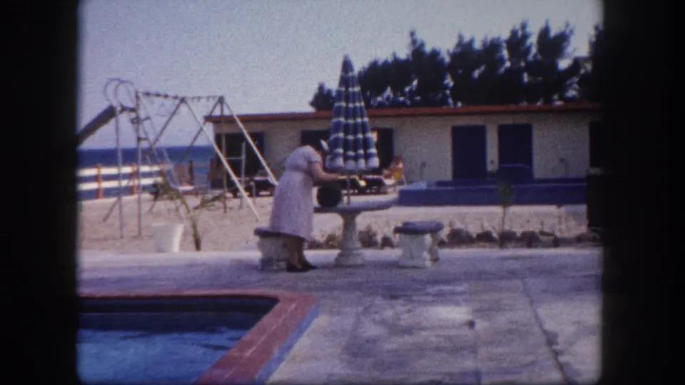 A woman leans over a concrete garden structure beside a Florida beach resort pool; a closed striped umbrella, swing-set frame, and single-story building with blue shutters are visible in the background