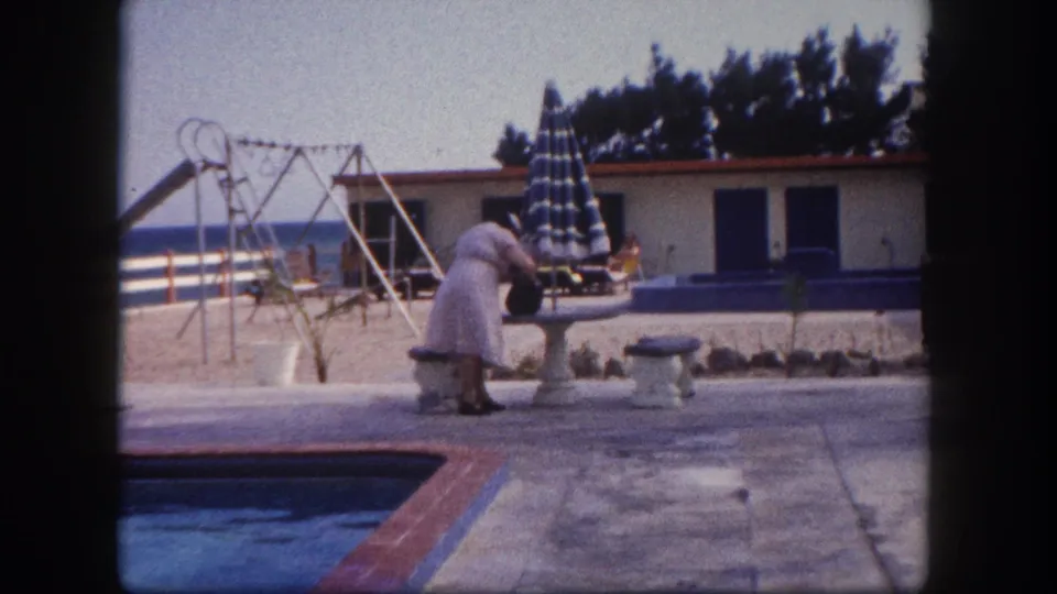 A woman in a 1960s dress stands near the edge of a Florida beach resort pool; a striped umbrella, swing-set frame, low white building with blue shutters, and ocean are visible in the background