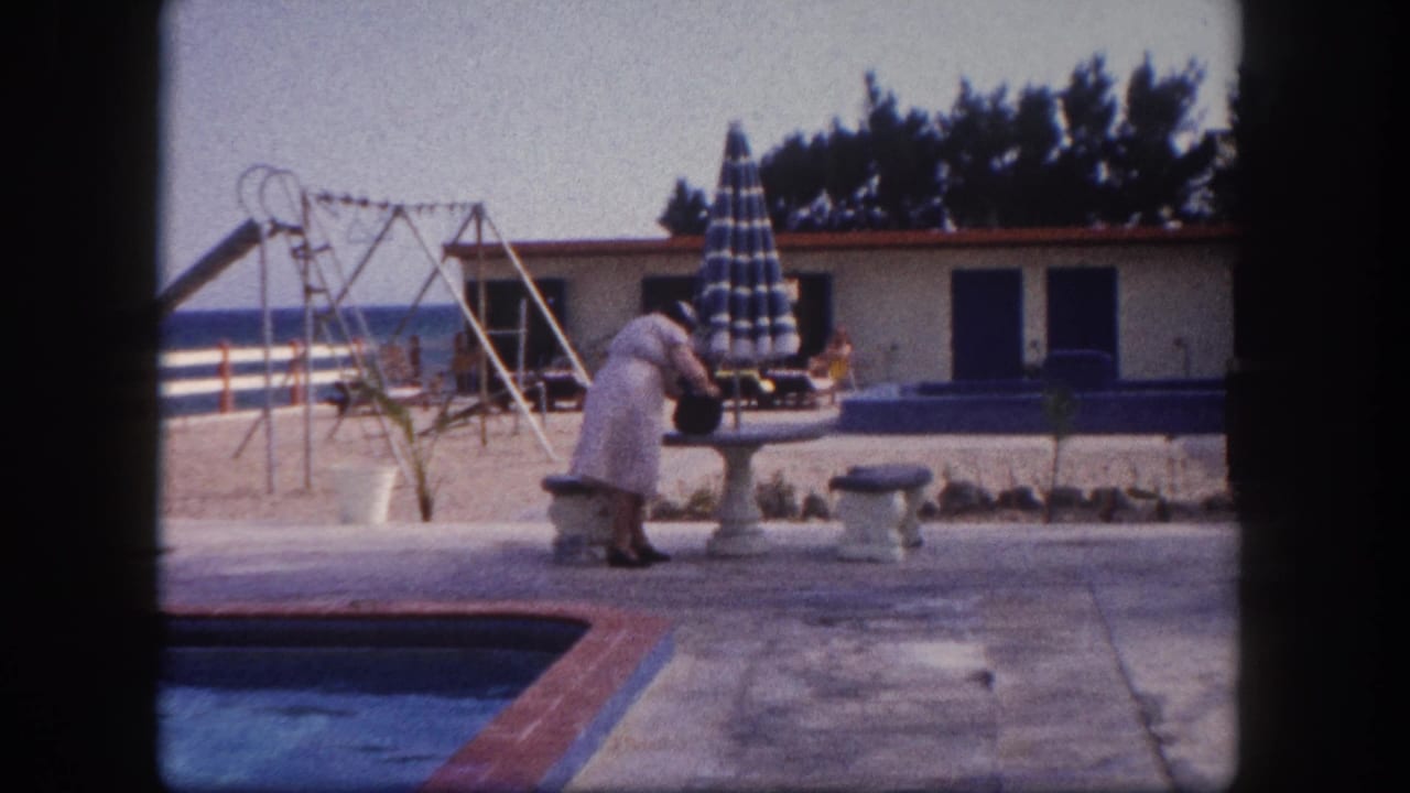 Elderly woman bending to retrieve a dark bag from a stone table at a Florida resort poolside, with furled striped umbrella and playground equipment visible, circa 1960.