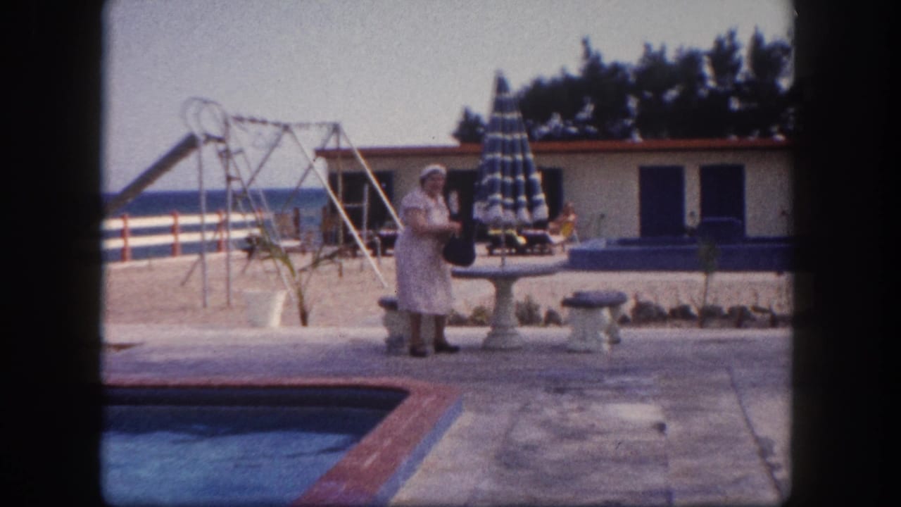 Elderly woman standing with dark handbag at a resort poolside, with striped umbrella, playground equipment, blue-shuttered building, and ocean horizon visible, Florida circa 1960.
