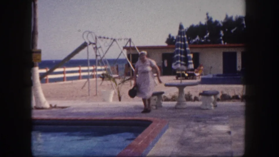 Elderly woman walking along a pool deck toward camera carrying a dark handbag, with striped umbrella, playground slide, and flat-roofed building behind her, Florida resort circa 1960.