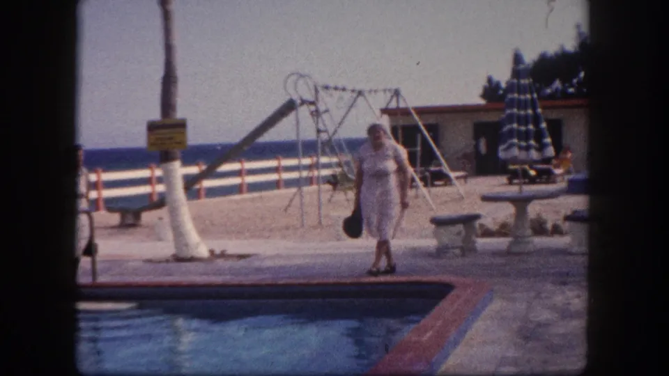 Elderly woman approaching camera on a Florida resort poolside patio, carrying a dark handbag, with palm tree, swing set, building, and ocean horizon visible, circa 1960.