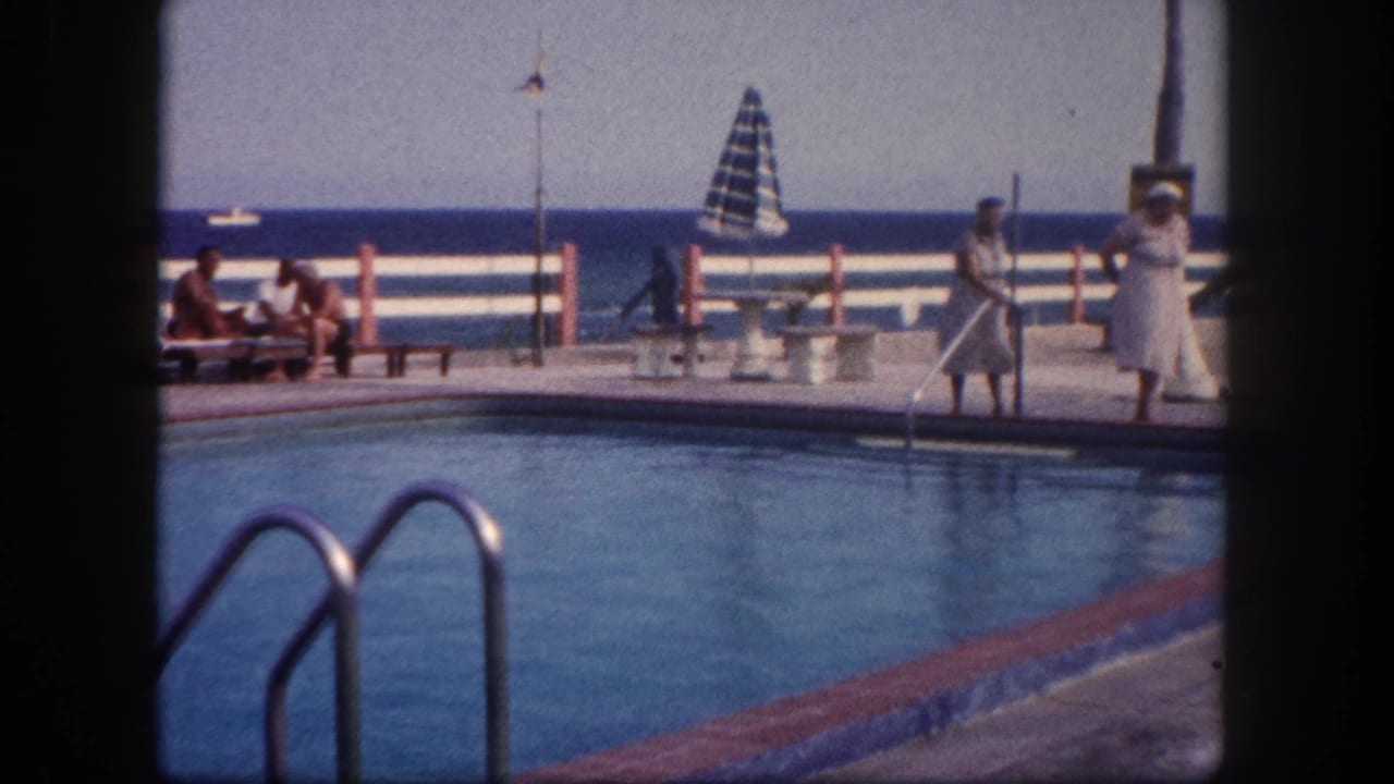 8mm home movie frame circa 1960, Florida — outdoor poolside at a marina or club, figures near waterfront railing, sailboat on open water beyond.