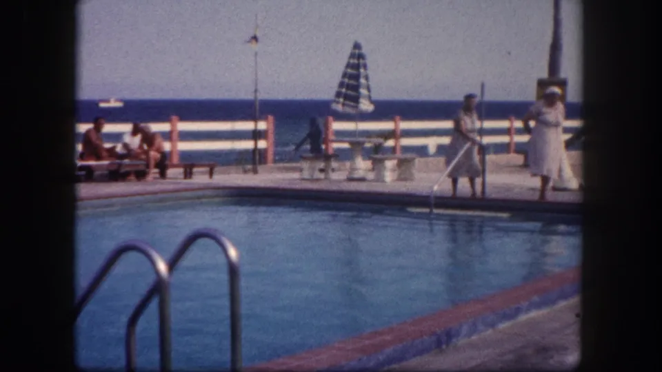 8mm home movie frame circa 1960, Florida — outdoor poolside at a marina or club, figures near waterfront railing, sailboat on open water beyond.