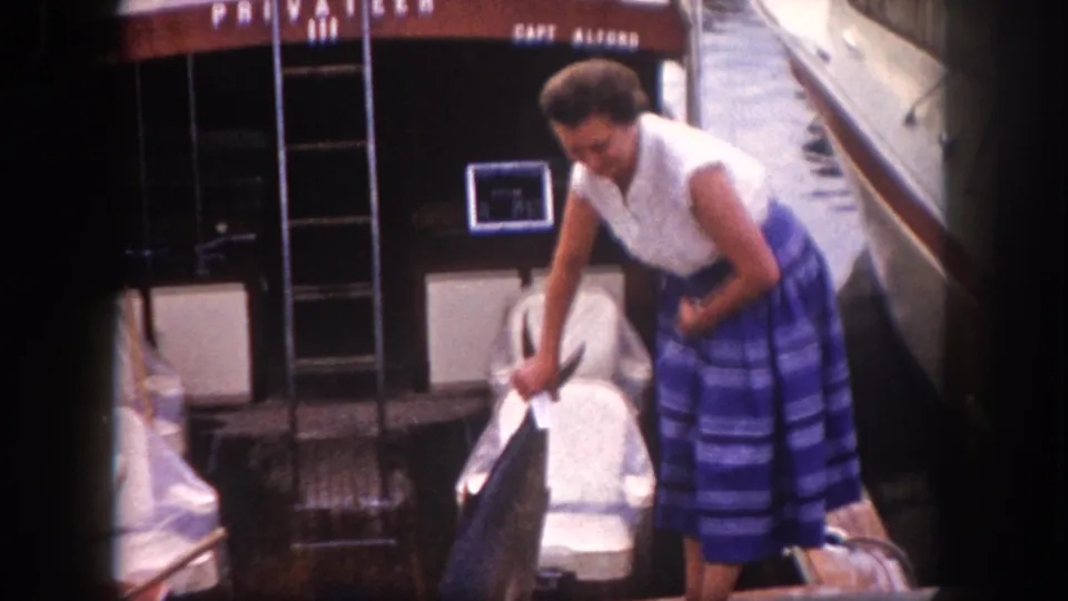 Early 1960s 8mm frame, Tampa Florida — woman leans toward camera smiling with large fish; dock furniture and PRIVATEER III stern visible in background.