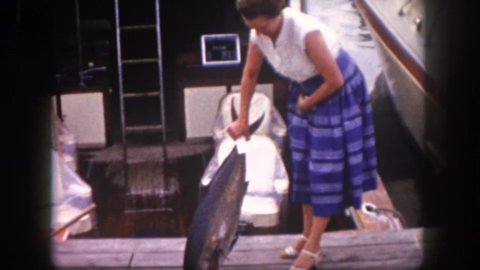 Early 1960s 8mm frame, Tampa Florida — woman holds large game fish at dock; PRIVATEER III stern and second moored vessel visible; heeled sandals in frame.