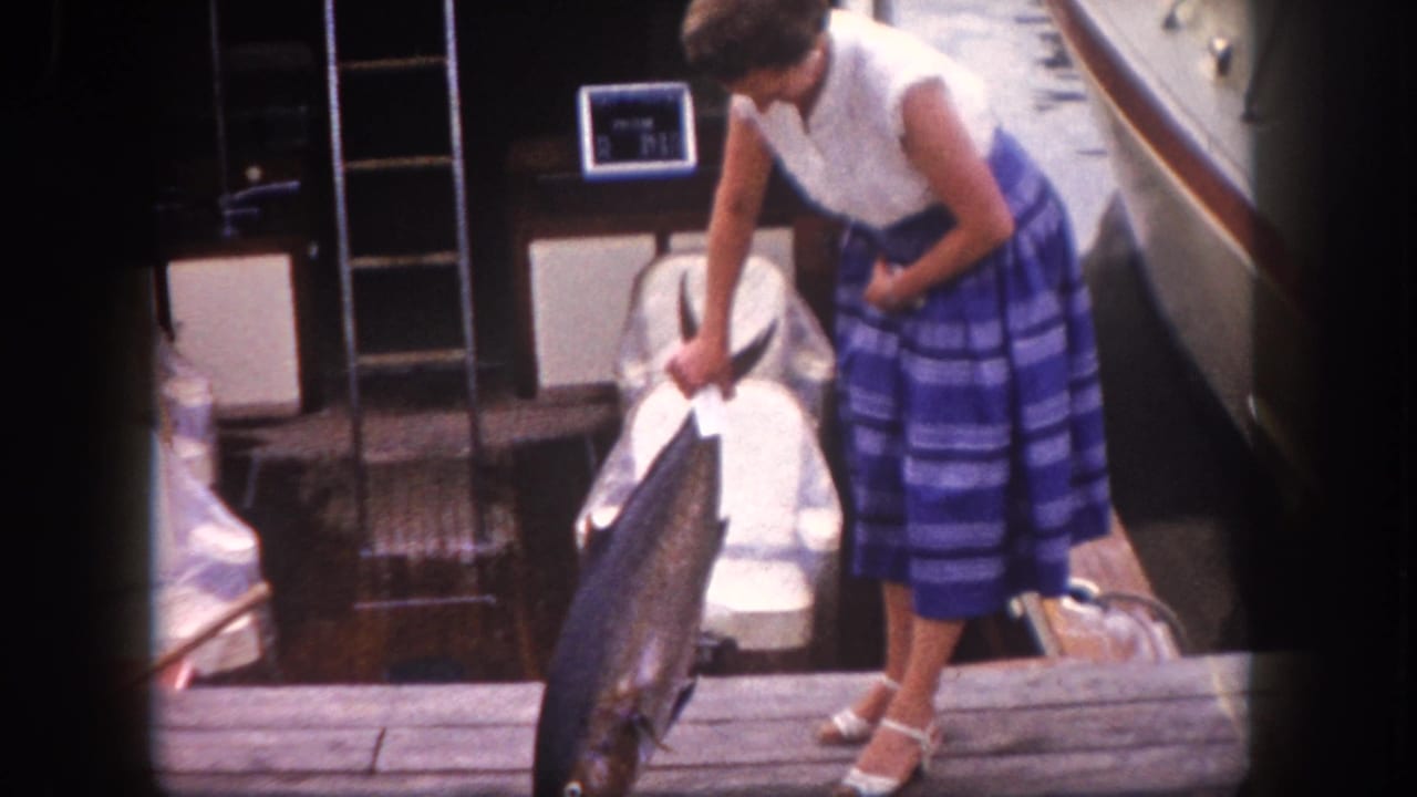 Woman in white sleeveless top and blue striped skirt stands on a dock looking down at a large fish she is holding.