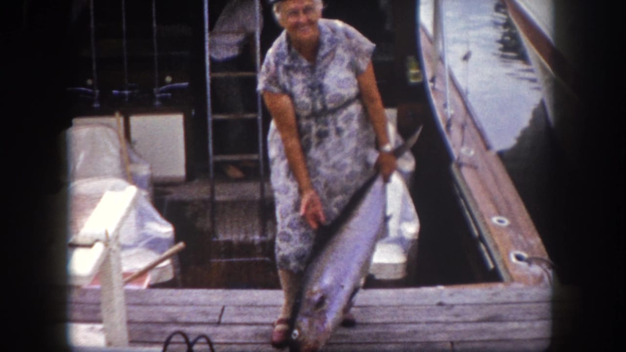 Older woman in patterned dress and boating cap holds a large fish upright on a wooden dock beside a motorboat.
