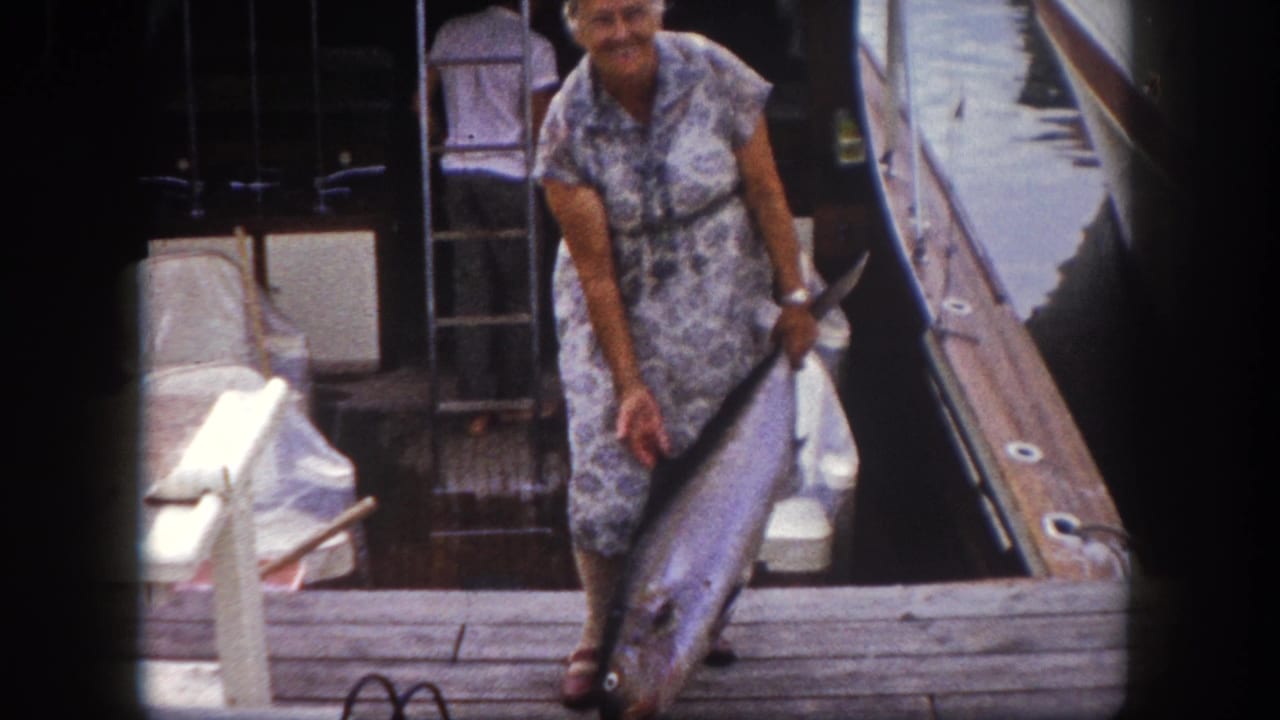 Woman in floral shift dress and white boating cap holds a large sport fish on a marina dock, Florida, circa 1960.
