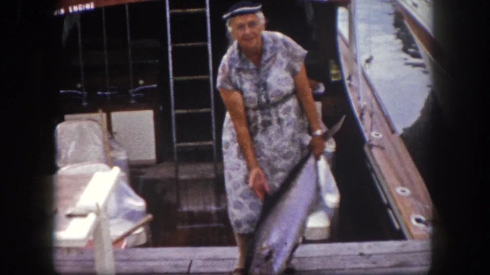 Woman displays a large sport fish on a wooden dock beside a motorboat with partial boat lettering visible, Florida marina, circa 1960.