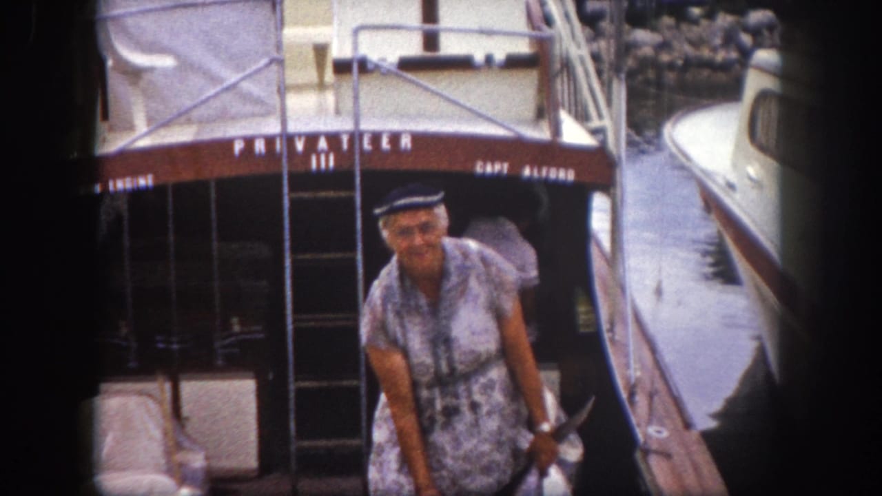 Older woman in patterned dress and nautical cap standing on deck of boat named Privateer III at a marina.