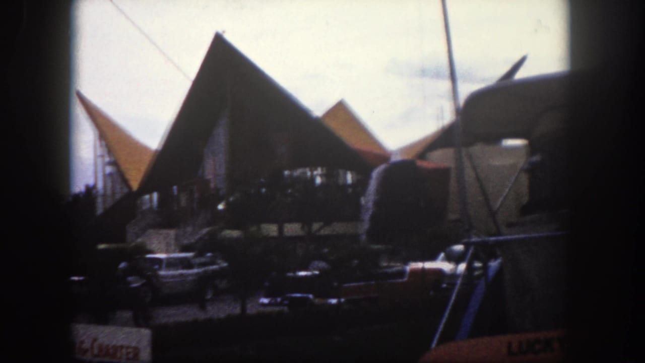 Angular pointed rooftop canopies and early-1960s parked cars on a Florida commercial street, viewed through a vehicle window.
