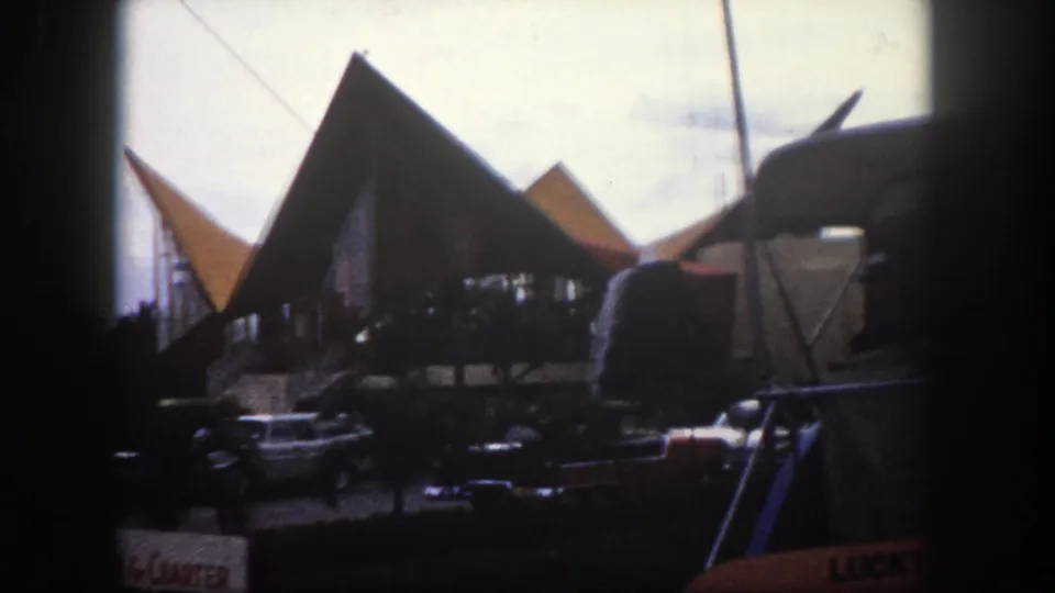 Pointed A-frame or tent-style commercial canopy structures and era-appropriate American cars on a Tampa, Florida street, circa 1960, on 8mm color film.