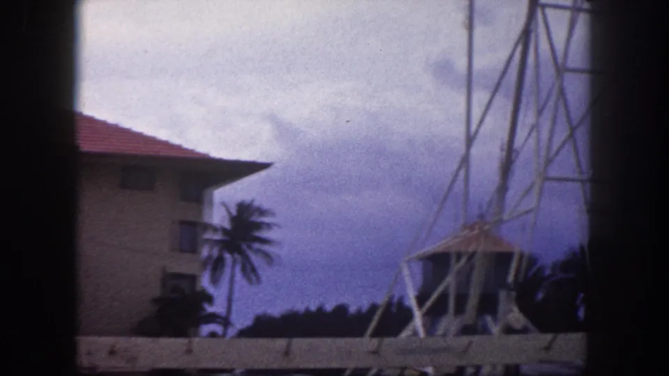 8mm home movie frame, Tampa, Florida, circa 1960: metal lattice tower with palm trees and a red-tiled building against a blue-toned overcast sky.
