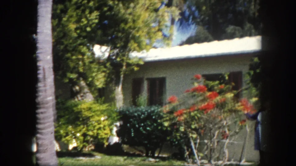 8mm home movie frame, Tampa, Florida, circa 1960: woman in blue dress stands beside a flowering garden in front of a single-story teal house.