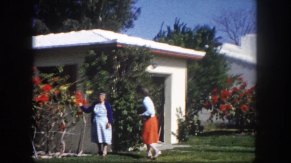 Two women stand together beside a large red flowering bush, white stucco ranch house visible behind them, circa 1960 Tampa, Florida, 8mm home movie.