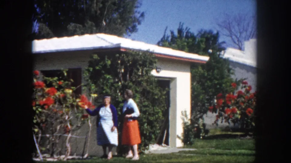 Two women beside a large red flowering bush facing the camera, white stucco house behind, circa 1960 Tampa, Florida, 8mm home movie.