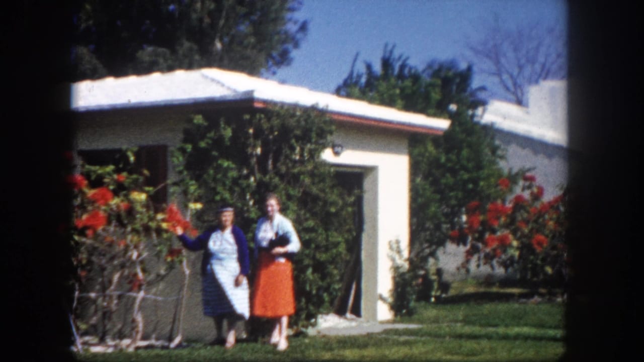 Two women — one in a blue cardigan and striped dress, one in an orange skirt — pose beside a white flat-roofed stucco house with red flowering shrubs, Tampa Florida, circa 1960.