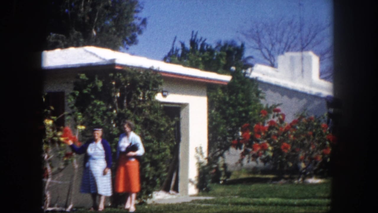 Two women in colorful 1960s clothing stand together in a sunny garden in front of a white stucco house with red flowering bushes.