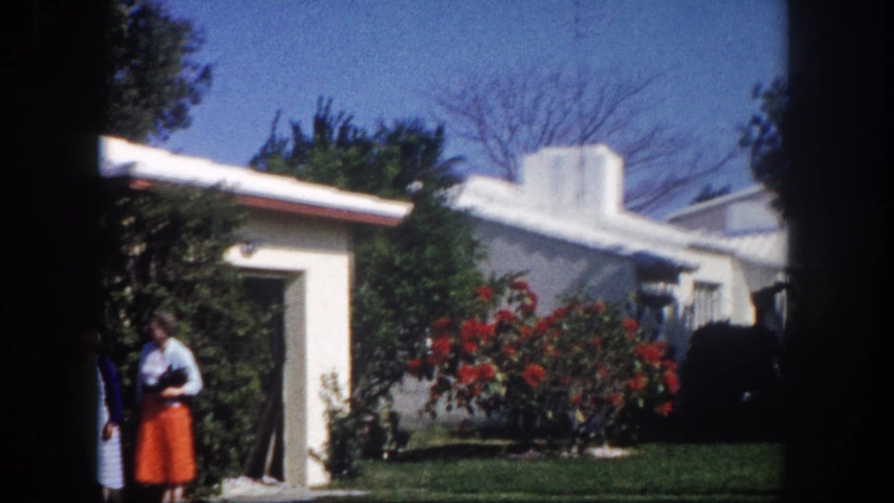 Wider home-movie frame showing a white flat-roofed stucco ranch house with red flowering shrub, green lawn, and bare trees against a blue sky; one woman partially visible, Tampa Florida circa 1960.